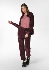 Completo tuta in velour borgogna composto da una giacca con zip e pantaloni abbinati, abbinato a una t-shirt rosa e scarpe da ginnastica nere. Finitura testurizzata.