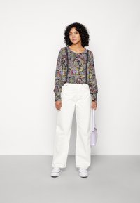 Top de manga longa com padrão floral e punhos elásticos, combinado com calças largas brancas. Segura uma bolsa lilás clara e usa sapatos brancos.