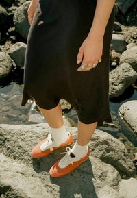 Oranžiniai ir balti sportbačiai su tekstūruotais paviršiais, baltais raišteliais ir žaliais akcentais; dėvimi su baltais nėriniais iki kulkšnies ir juoda suknele. Fone matyti akmenys.