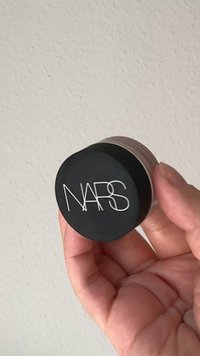 Pot noir rond avec une finition mate, arborant le logo blanc "NARS" sur le dessus. Base transparente avec une texture lisse, tenu dans une main.