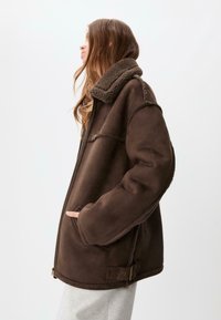 Giubbotto in pelliccia sintetica marrone con colletto morbido, chiusura con zip, tasche laterali e vestibilità rilassata. Presenta dettagli di cucitura a vista.