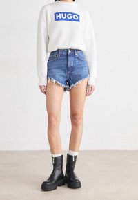 Maglione bianco a coste con logo blu "HUGO", pantaloni denim a vita alta con orlo sfilacciato, e stivaletti neri alla caviglia con zip laterali.