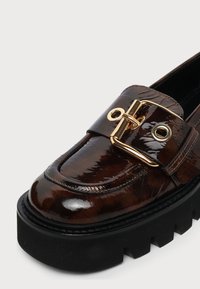 Mocassin marron foncé brillant avec une semelle noire épaisse et une grande boucle dorée sur le dessus.
