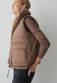 Braune Daunenweste mit hohem Kragen, Frontreißverschluss und einer aufgenähten Tasche. Weiche Fütterung und Kordelzug-Detail am Saum. Getragen über einem braunen Langarmshirt.