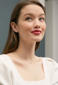 Model nosi bijelu teksturiranu bluzu s balon rukavima, srebrnu ogrlicu s malim privjeskom i naušnice koje izgledaju poput dijamanata.