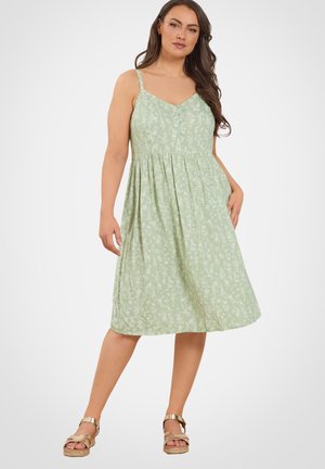 Kobieta ubrana w bezrękawową, jasnozieloną, kwiatową sukienkę midi z guzikami, stoi z jedną ręką w kieszeni sukienki i złotymi sandałami.