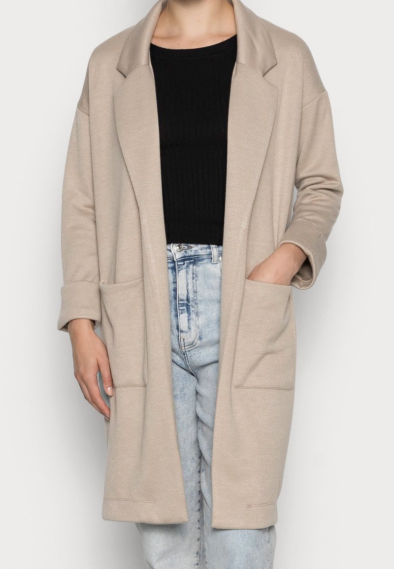 Beige long jacket with rolled sleeves and front pockets worn over a black ribbed top and light blue high-waisted jeans.