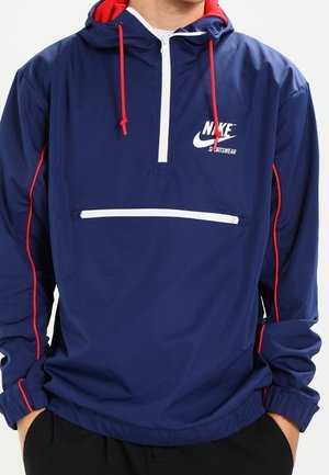 Homme portant un coupe-vent Nike bleu marine avec des cordons rouges et un liseré, fermeture éclair frontale mi-longue, et une poche zippée blanche horizontale.