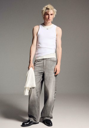 Mladi oseba z razmršenimi blond lasmi, oblečena v belo majico brez rokavov, ohlapne sive kavbojke, črne čevlje, drži bel kos blaga na sivem ozadju.