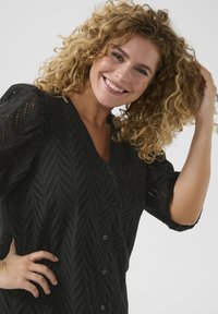 Haut noir à boutons avec une texture à motif de feuilles et des manches courtes bouffantes, mettant en valeur un décolleté en V profond et des découpes décoratives.