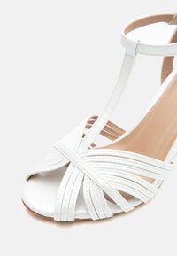 Λευκό πέδιλο με ψηλό τακούνι, ανοιχτό δάχτυλο, με σταυρωτά λουράκια και λουράκι στον αστράγαλο, σε ανοιχτό φόντο.