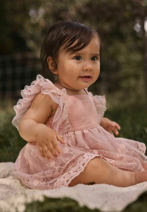 Bambina seduta su una coperta all'aperto, indossa un vestito di pizzo rosa e guarda leggermente di lato con un'espressione calma.