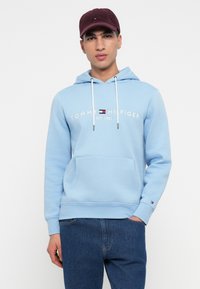 Νεαρός άνδρας φορεί ελαφρώς μπλε φούτερ Tommy Hilfiger, σκούρα μπλε τζιν και μαραντον καπέλο, ενώ στέκεται μπροστά σε λευκό φόντο.