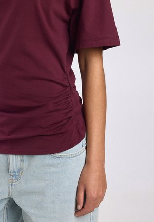 Donkerrode T-shirt met korte mouwen en zijsamenstelling, gedragen met lichtblauwe spijkerbroek, arm en hand zichtbaar tegen een witte achtergrond.