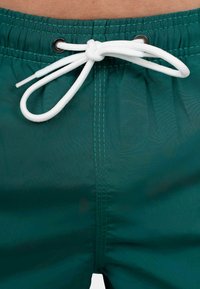 Costumi da bagno turchesi, con un laccetto bianco e occhiello in metallo, presentano un materiale liscio e leggero e cuciture a contrasto.
