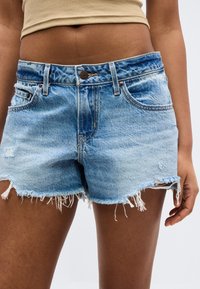 Gros plan d'une personne portant un short en jean bleu clair usé et un top court beige, montrant le ventre et le haut des cuisses.