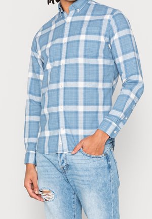 Homme portant une chemise à carreaux larges bleu clair et blanc à manches longues et un jean bleu clair délavé, main dans la poche.