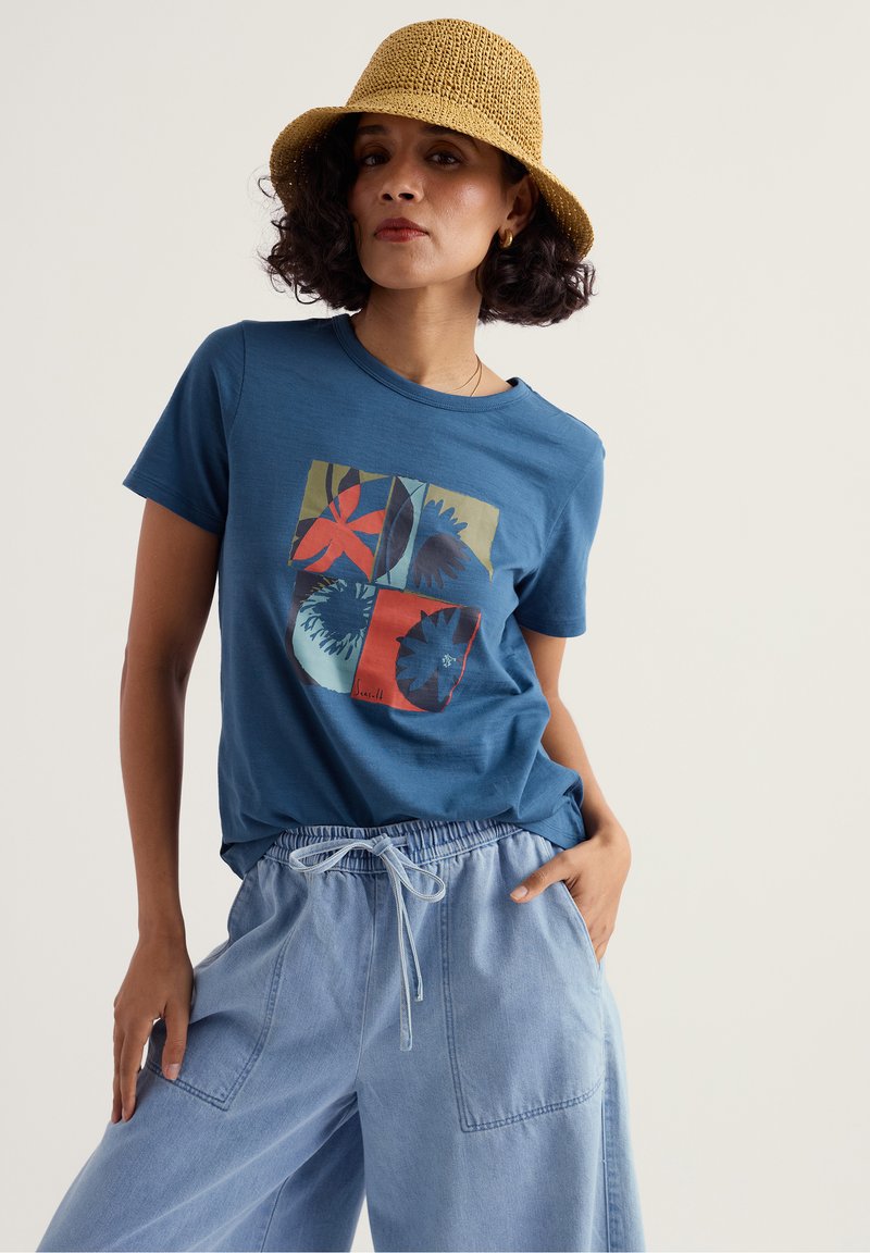 Frau mit blauem T-Shirt mit Grafikdruck, hellblauen Hose mit Kordelzug, gelbem gewebtem Hut und goldenen Creolen, die mit einer Hand in der Tasche steht.