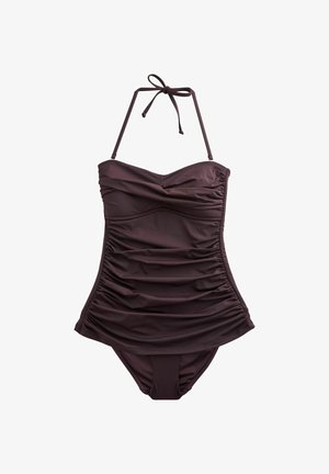 Jeune femme aux longs cheveux foncés portant un maillot de bain une pièce sombre, marchant sur une plage rocheuse ensoleillée près de l'eau.