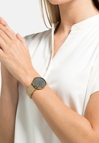 Goldener Mesh-Armbanduhr an einer Hand mit einem dunkelgrauen Zifferblatt. Die Uhr verfügt über Stundenmarkierungen und ein minimalistisches Design und wird mit einer weißen Bluse kombiniert.
