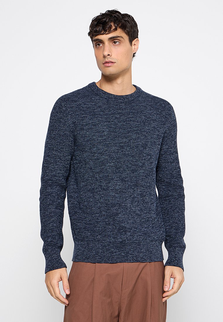 Junger Mann mit dunklem, lockigem Haar, der einen strukturierten, marineblauen Pullover und braune Hosen trägt, steht vor einem schlichten weißen Hintergrund.