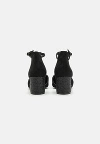 Zapatos de tacón alto de ante negro con parte trasera abierta, correas ajustables para el tobillo y tacones de bloque texturizados en negro.