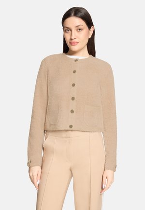 Femme portant un cardigan beige à boutons en peluche et un pantalon beige taille haute, debout avec les bras détendus sur un fond uni.