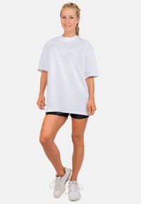 Overmaat wit t-shirt met korte mouwen, voorzien van een cursief logo op de voorkant. Ggedragen met zwarte shorts en witte sportschoenen.