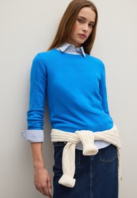 Blauwe wollen trui met een ronde hals, gedragen over een blauw-witte gestreepte overhemd, gecombineerd met een spijkerrok en een witte kabeltrui die om de taille is geknoopt.