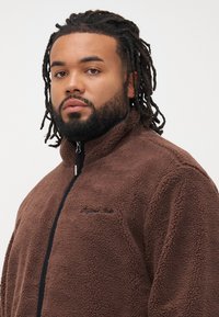 Homme avec une barbe et des dreadlocks portant une veste en polaire brun zippée sur un fond clair uni.