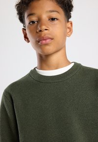 Maglione verde scuro con scollatura a girocollo a coste, realizzato in tessuto testurizzato, con colletto interno bianco. Primo piano della vista frontale.