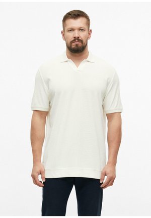 Mann mit kurzen Haaren und Bart, der ein schlichtes weißes kurzärmeliges Poloshirt und dunkle Hosen trägt, steht vor einem weißen Hintergrund.