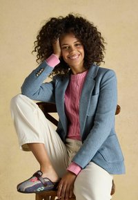 Femeie cu păr creț, zâmbitoare, așezată pe un scaun din lemn, purtând sacou albastru, pulover roz, pantaloni albi și pantofi slip-on cu model.