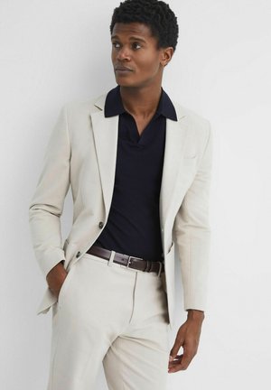 Traje beige claro con diseño de una sola botonadura, combinado con una camiseta polo azul marino y cinturón marrón. La textura suave y el ajuste a medida son evidentes.