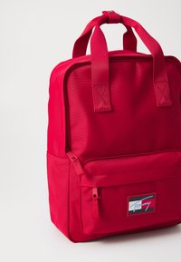 Sac à dos en tissu rouge avec deux poignées supérieures, une poche zippée à l'avant et un patch de logo. Il présente une forme rectangulaire et une surface texturée.