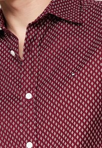 Bordeaux knoopshirt van een textuurstof, met een wit bloemenpatroon en een klassieke kraag, vergezeld van witte knopen.