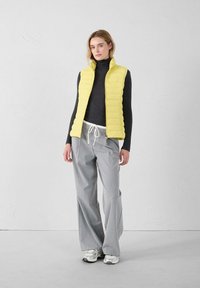 Chaleco amarillo acolchado sobre un cuello de tortuga negro, combinado con pantalones sueltos gris claro y zapatillas blancas. Textura suave, diseño minimalista.
