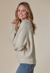 Hellbeiger Oversize-Pullover mit strukturierter Oberfläche, geripptem Saum und überschnittenen Schultern. Verfügt über einen kleinen Logo-Patch an der Seite.