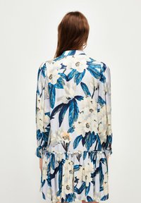 Blumenkleid mit langen Ärmeln, das ein blau-weißes Blumenmuster, einen gerafften Saum und eine glatte Stoffoberfläche aufweist.