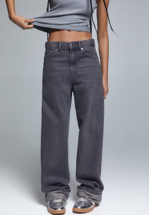 Person wearing loose dark gray jeans with rolled cuffs and silver sneakers, lifting a gray ribbed crop top, showing midriff.