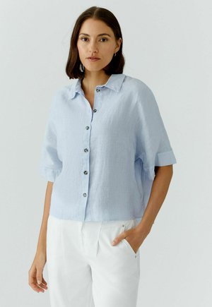 Vrouw met donker haar draagt een lichtblauw kortermig overhemd met knopen en witte broek, staand met één hand in de zak tegen een effen achtergrond.