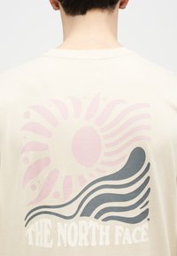 Οπίσθια όψη ατόμου που φοράει πουκάμισο εκρού με ροζ σχέδιο ηλιακής ακτινοβολίας και γκρι κυματιστό μοτίβο πάνω από το λογότυπο "The North Face" στην πλάτη.