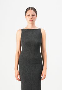 Ung kvinne med mørkt hår samlet i en hestehale, iført enermeløs, strukturert mørkegrå kjole, står foran en enkel hvit bakgrunn, vendt fremover.