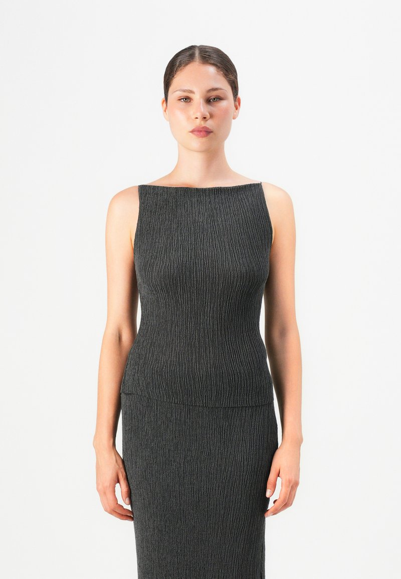 Ung kvinne med mørkt hår samlet i en hestehale, iført enermeløs, strukturert mørkegrå kjole, står foran en enkel hvit bakgrunn, vendt fremover.