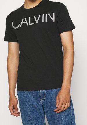 Zwart katoenen T-shirt met een grote zilveren "CALVIN"-opdruk over de borst, ronde hals en korte mouwen, gecombineerd met een blauwe spijkerbroek.