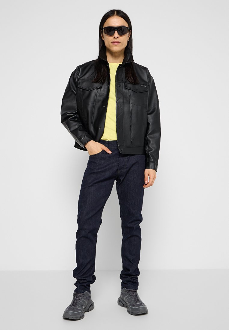 Giacca in finta pelle nera con chiusura a bottoni, abbinata a una camicia gialla e jeans blu scuri. Gli accessori includono occhiali da sole neri e sneakers grigie.