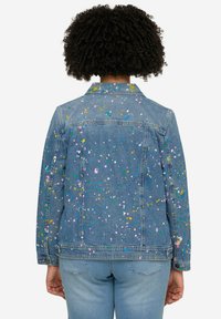 Veste en denim avec un motif de taches colorées. Dotée d'un col classique, de manches longues et d'une coupe droite. Tissu bleu avec une légère texture.