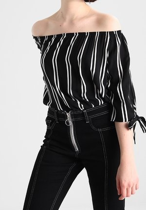 Femme portant un haut noir épaules dénudées à rayures blanches verticales et un pantalon noir taille haute avec une fermeture éclair visible à l'avant et des coutures blanches.