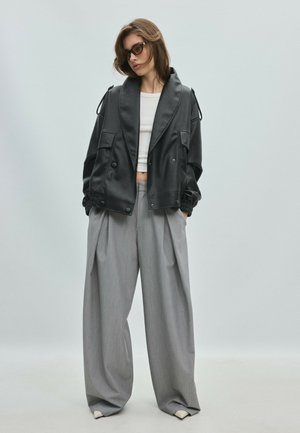 Žena má na sebe príliš veľkú čiernu koženú bundu, biely crop top, široké sivé nohavice a tmavé slnečné okuliare na jednoduchom svetlom pozadí.