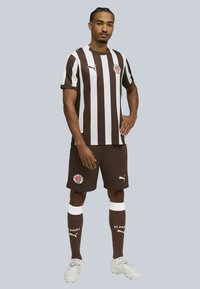 Brun og hvid stribet fodboldtrøje med matchende shorts. Har logo på brystet og shorts. Hvide accenter på sokker og fodtøj.
